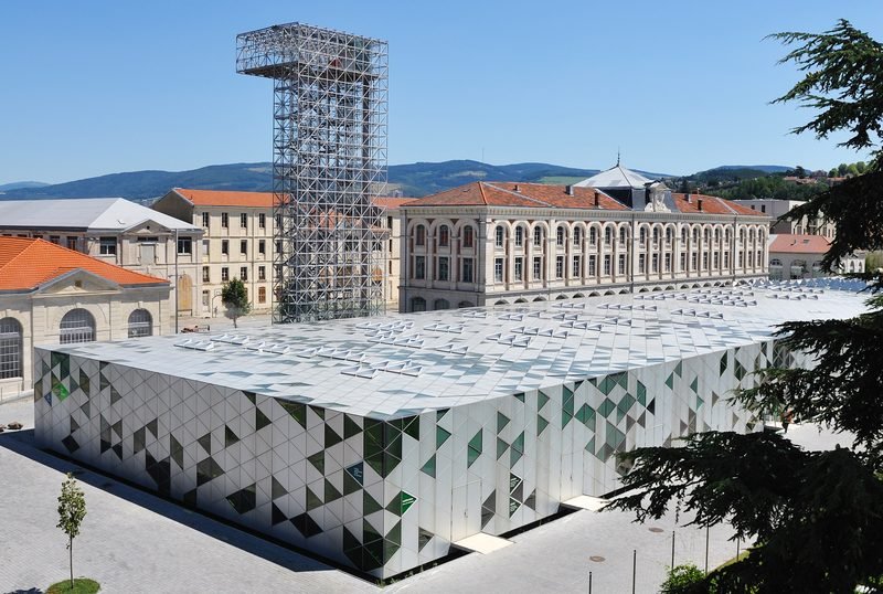 La Cité du Design à Saint-Étienne : bâtiment contemporain à façade triangulée, tour métallique et anciens bâtiments de la Manufacture, avec les collines en arrière-plan.